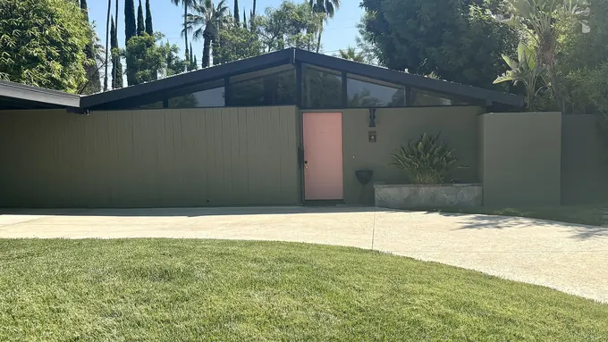 Low mid-century house with a pink front door behind a wide lawn and palms